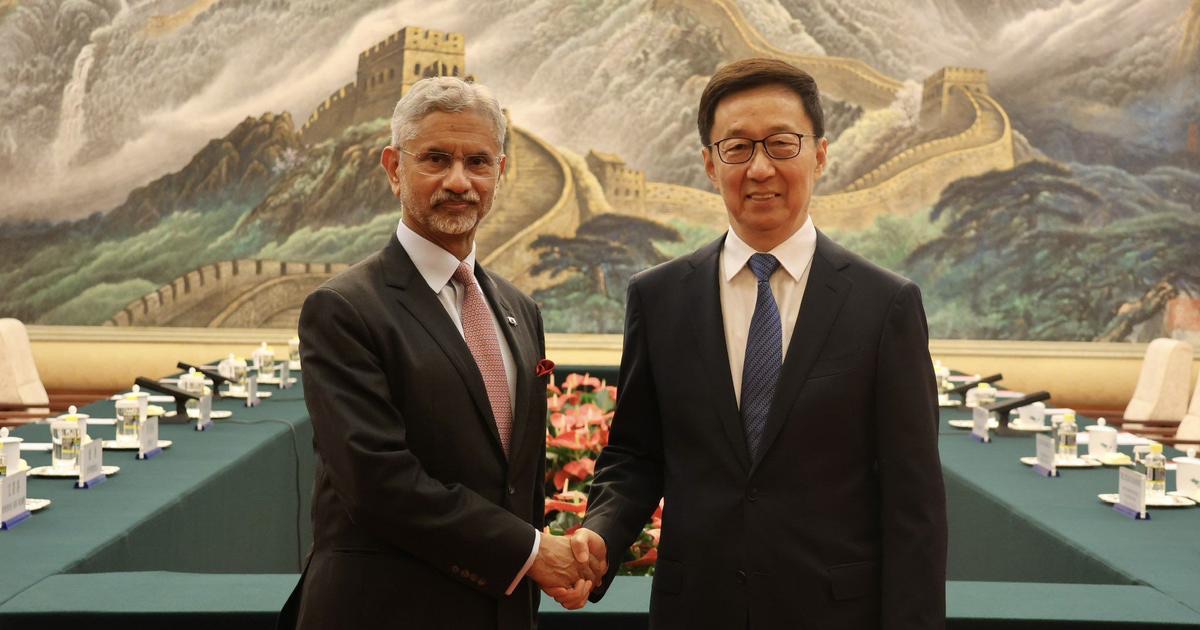Dr. S. Jaishankar shaking hands with a senior Chinese leader in a meeting hall with a large painting of the Great Wall