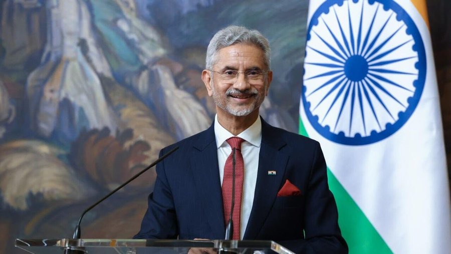 Dr. S. Jaishankar smiling at a lectern with microphones, the Indian flag behind him and a large painting to the left